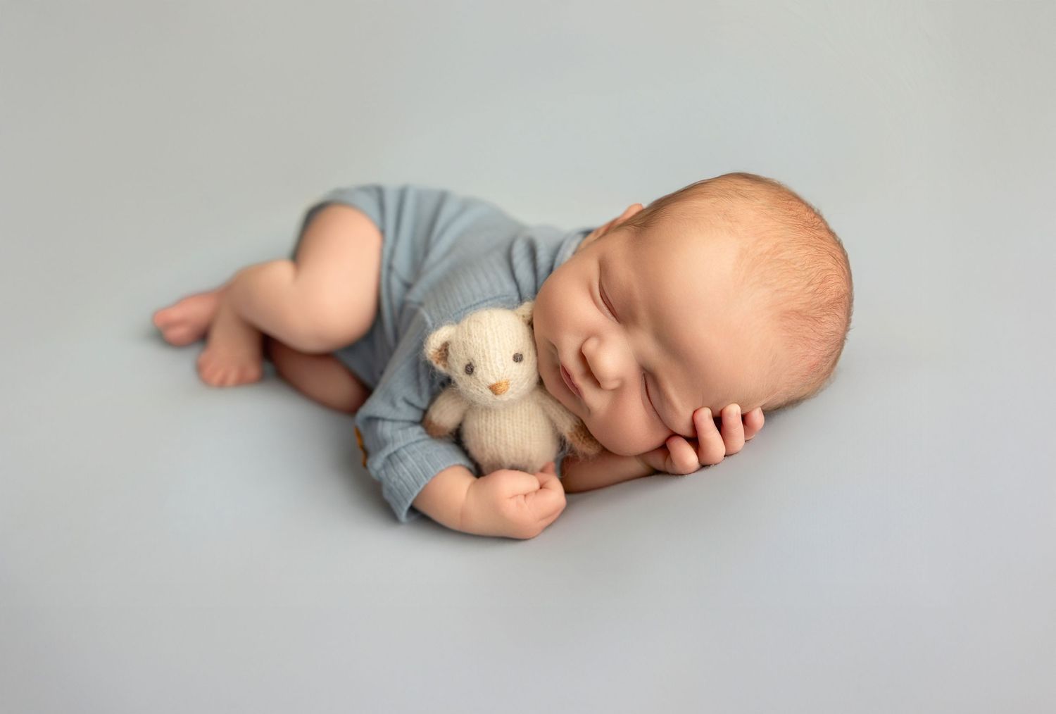 Sleeping newborn in gray onesie cuddling small teddy bear on light gray background.