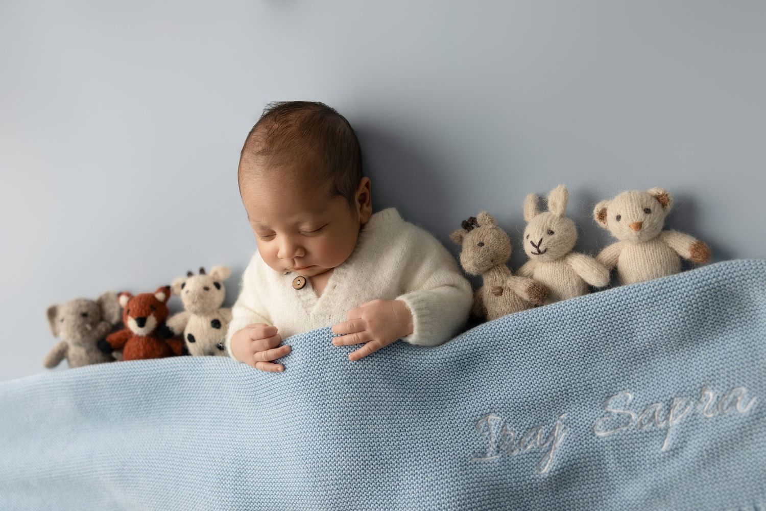 Sleeping baby in white sweater lying on blue blanket with collection of plush teddy bears in background.