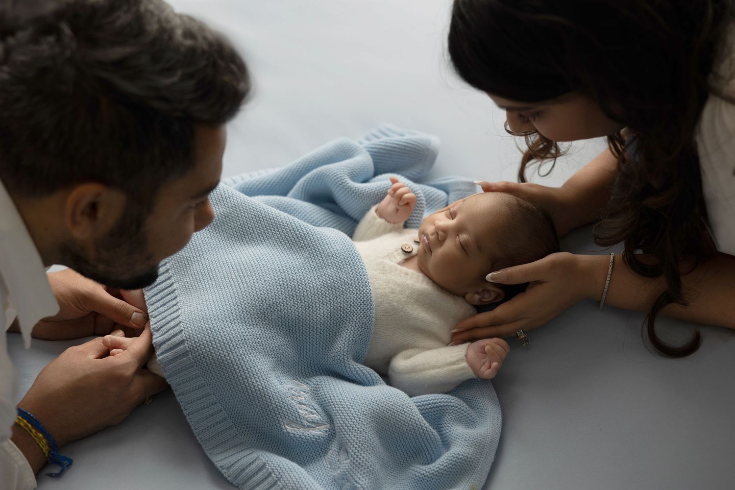 Baby wearing light blue sweater lies between two adults during an intimate family moment.