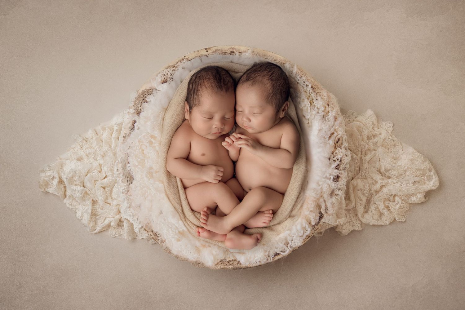 Two sleeping newborn babies nestled together in a cream-colored lace bowl on neutral backdrop.