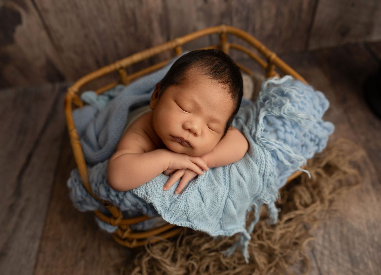A sleeping newborn baby wrapped in soft blue fabric lies peacefully in a rustic wooden setting.