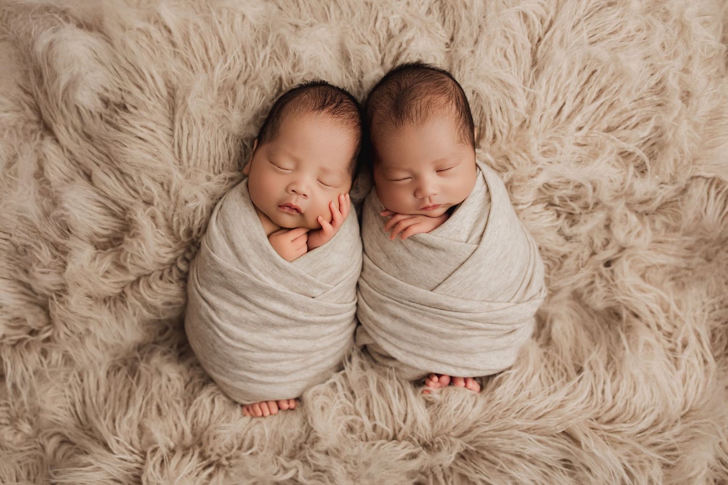 Newborn twins sleeping peacefully together on soft beige fur blanket in professional photography studio setting.