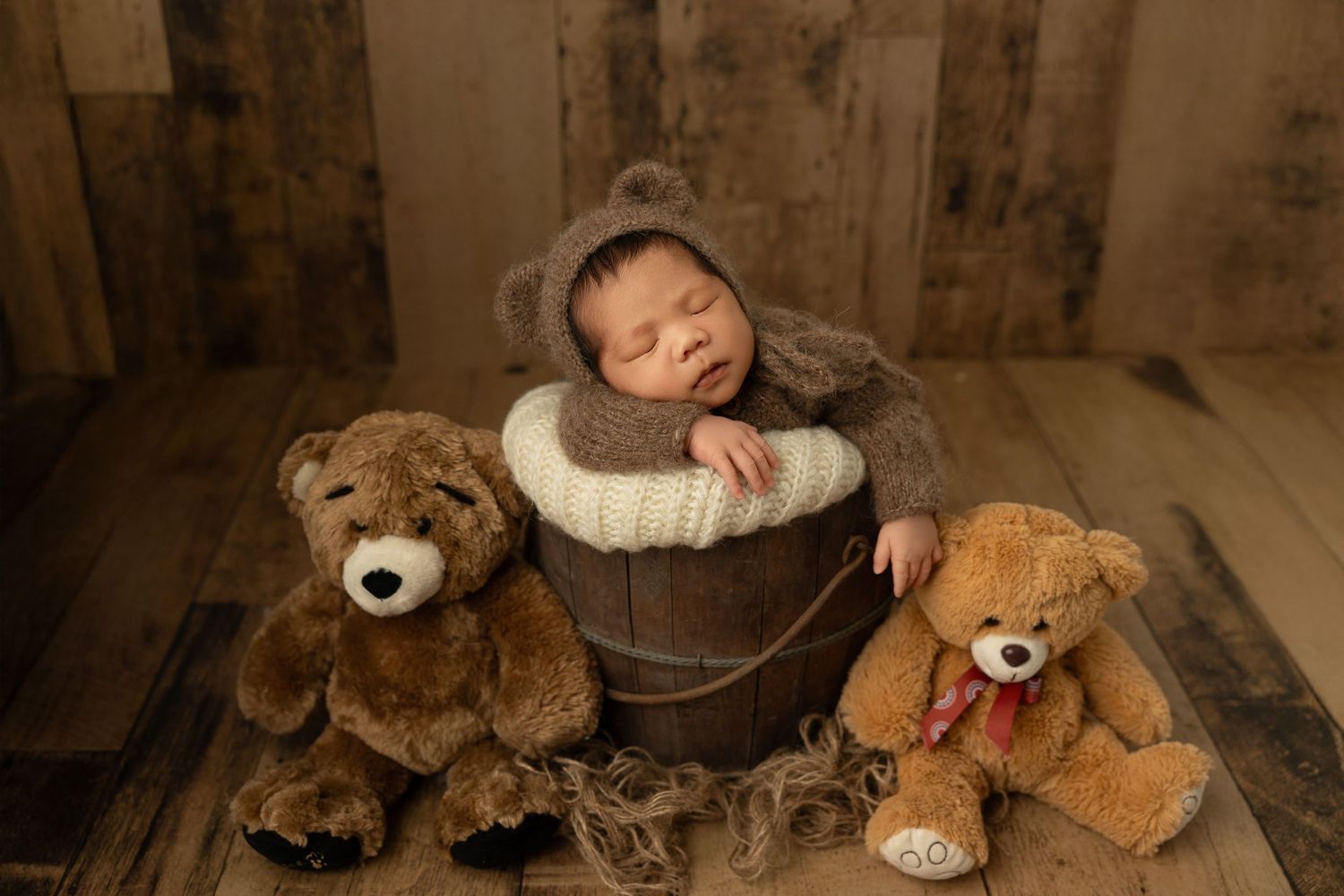 Baby rests in wooden bucket with two teddy bears on rustic wooden floor during newborn photography session.