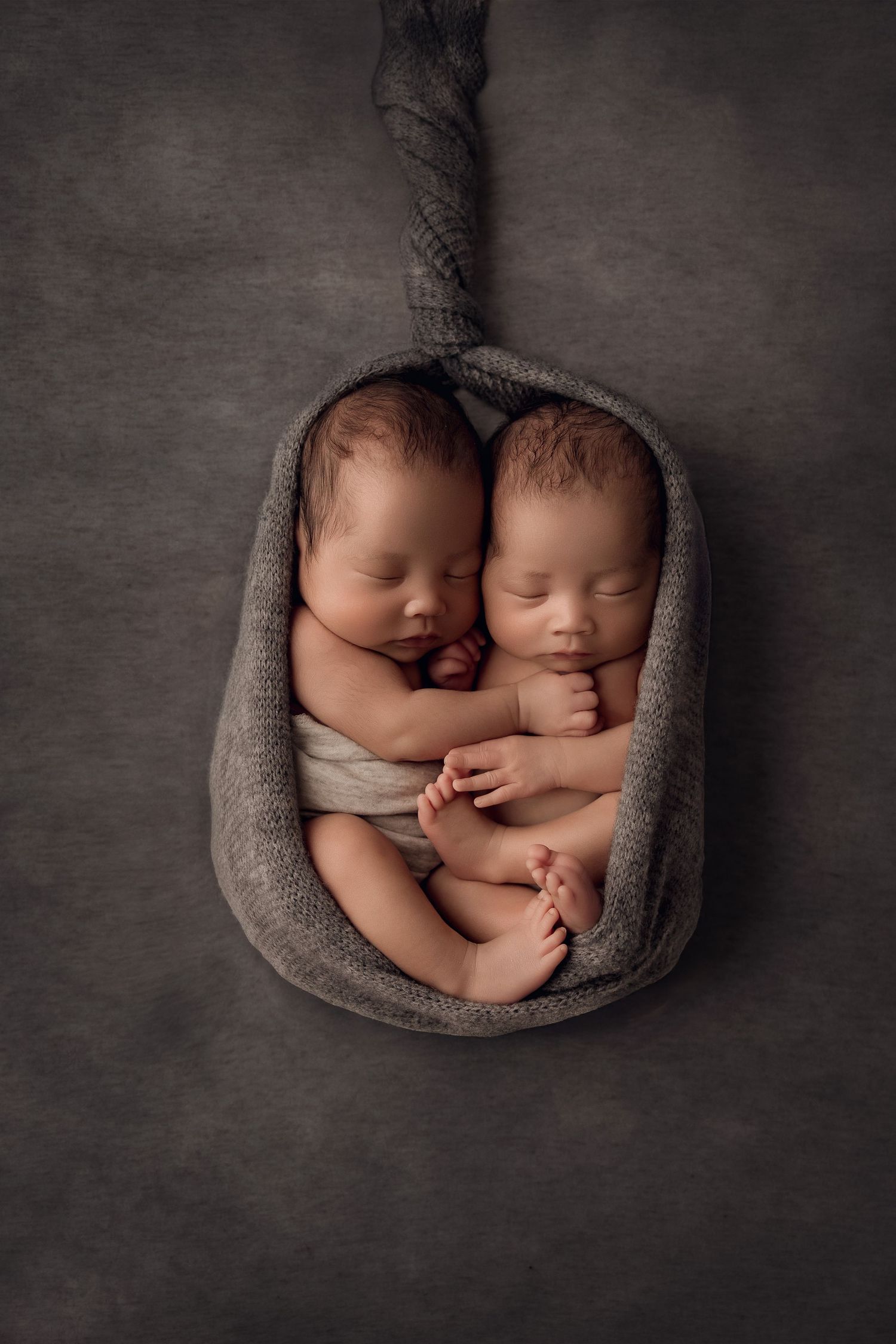 Peaceful sleeping newborn twins nestled together in a cozy gray knit wrap on dark background.