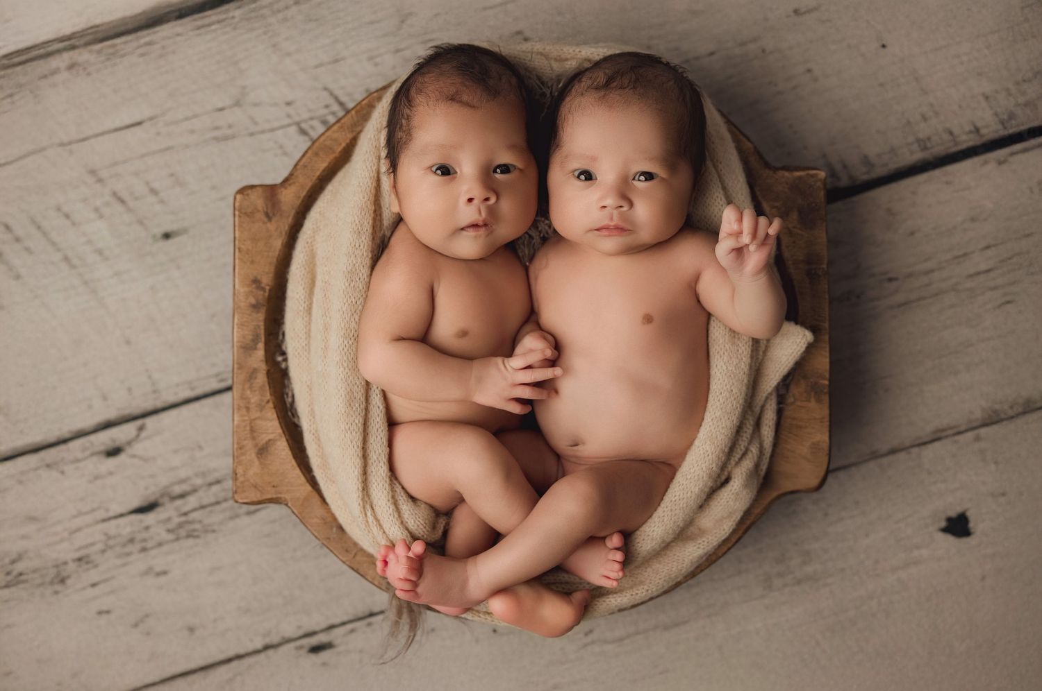 Twin babies nestled together in a round basket on rustic wooden floor during professional photo session.