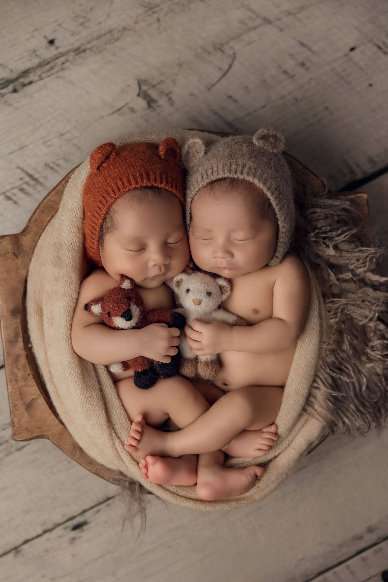Two sleeping newborns wearing knit hats cuddle together in a cozy beige blanket during a professional photo session.