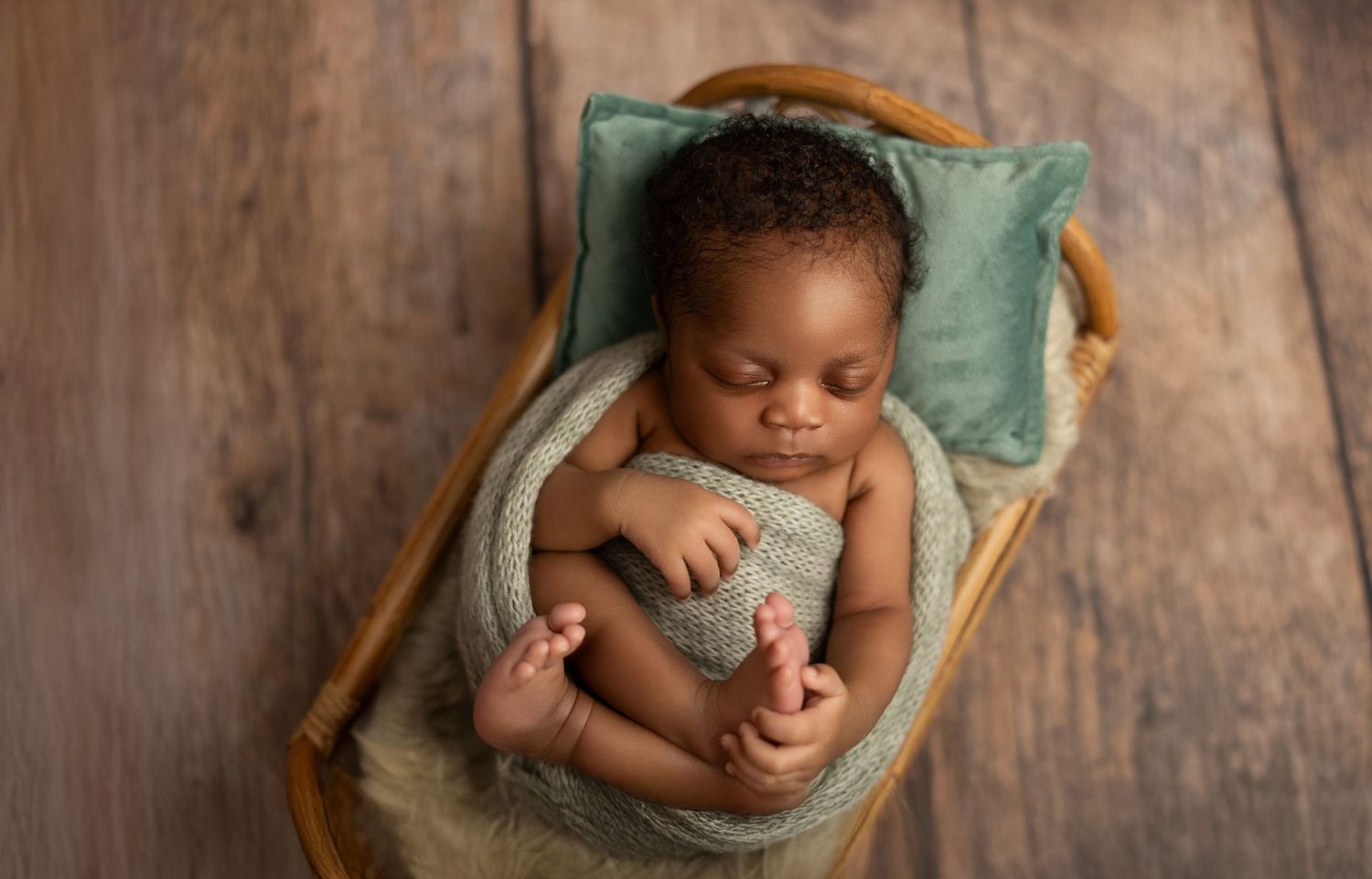 Sleeping newborn baby lying in a wooden basket with a mint green pillow on a wooden floor.