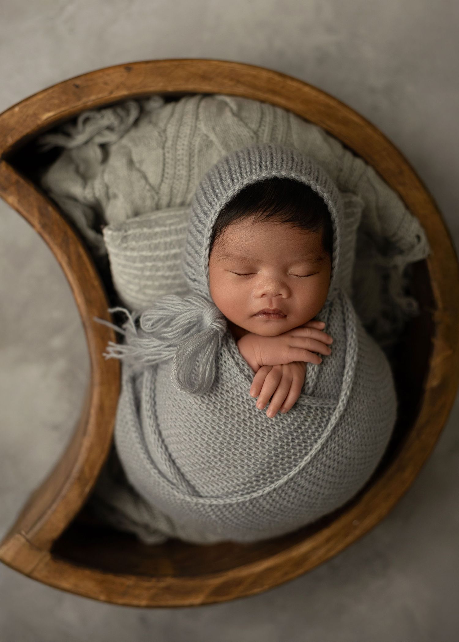 Sleeping newborn wrapped in soft gray knit swaddle lying in wooden bowl on neutral backdrop.