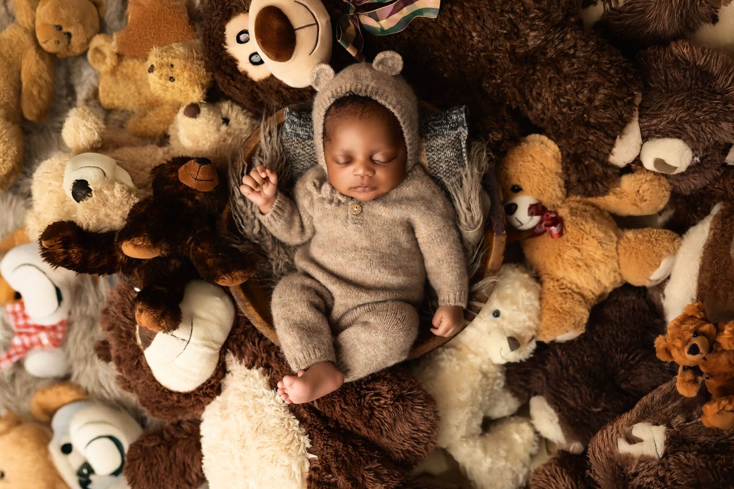 Newborn baby resting in grey outfit surrounded by plush teddy bears and stuffed animals.
