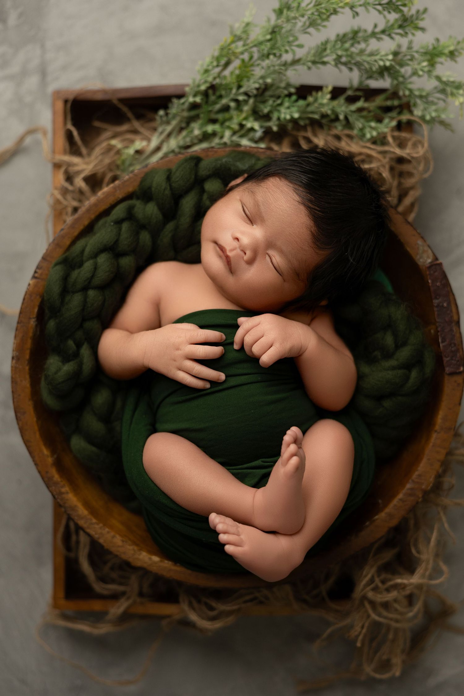 Newborn baby sleeping peacefully in wooden bowl with green wrap and natural decorative elements.