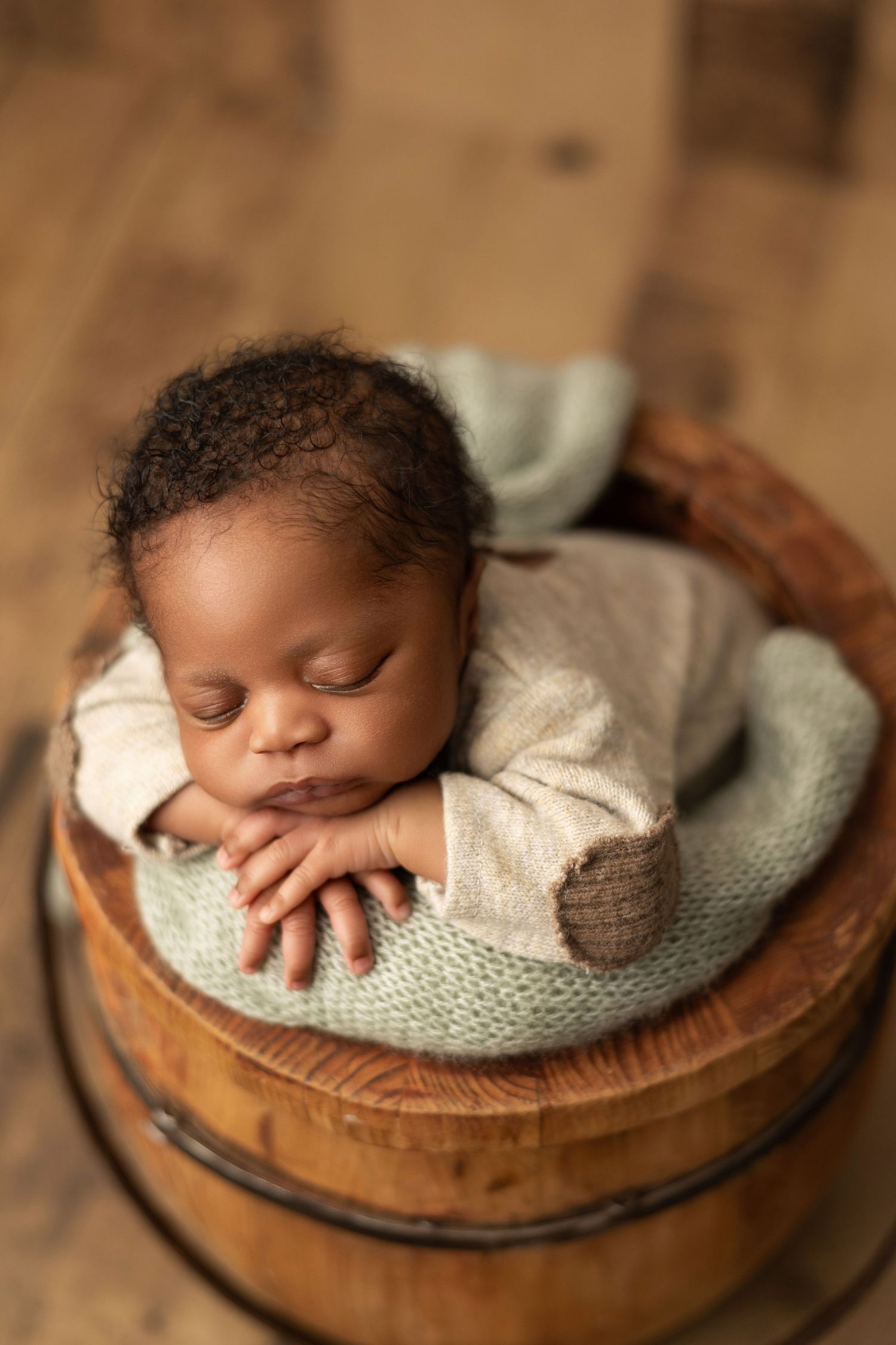 A sleeping newborn baby wrapped in a soft gray knit blanket rests peacefully in a wooden bowl.