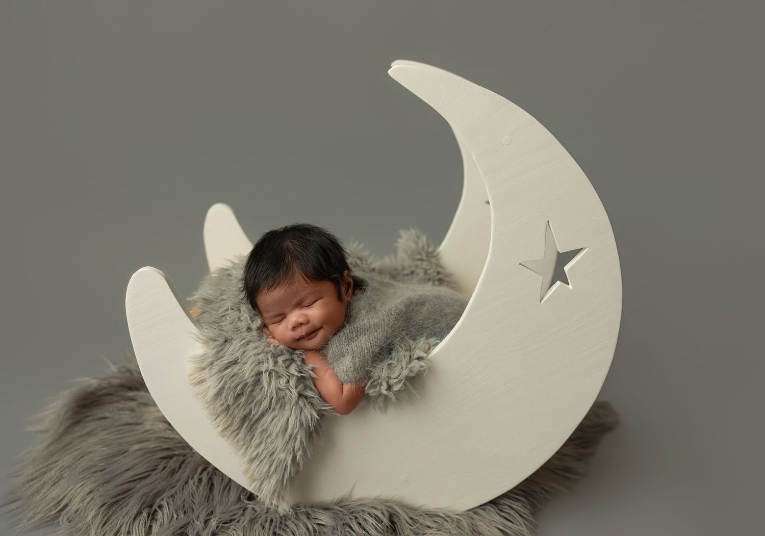 Sleeping newborn nestled in soft gray fur on a decorative white crescent moon prop with star detail.