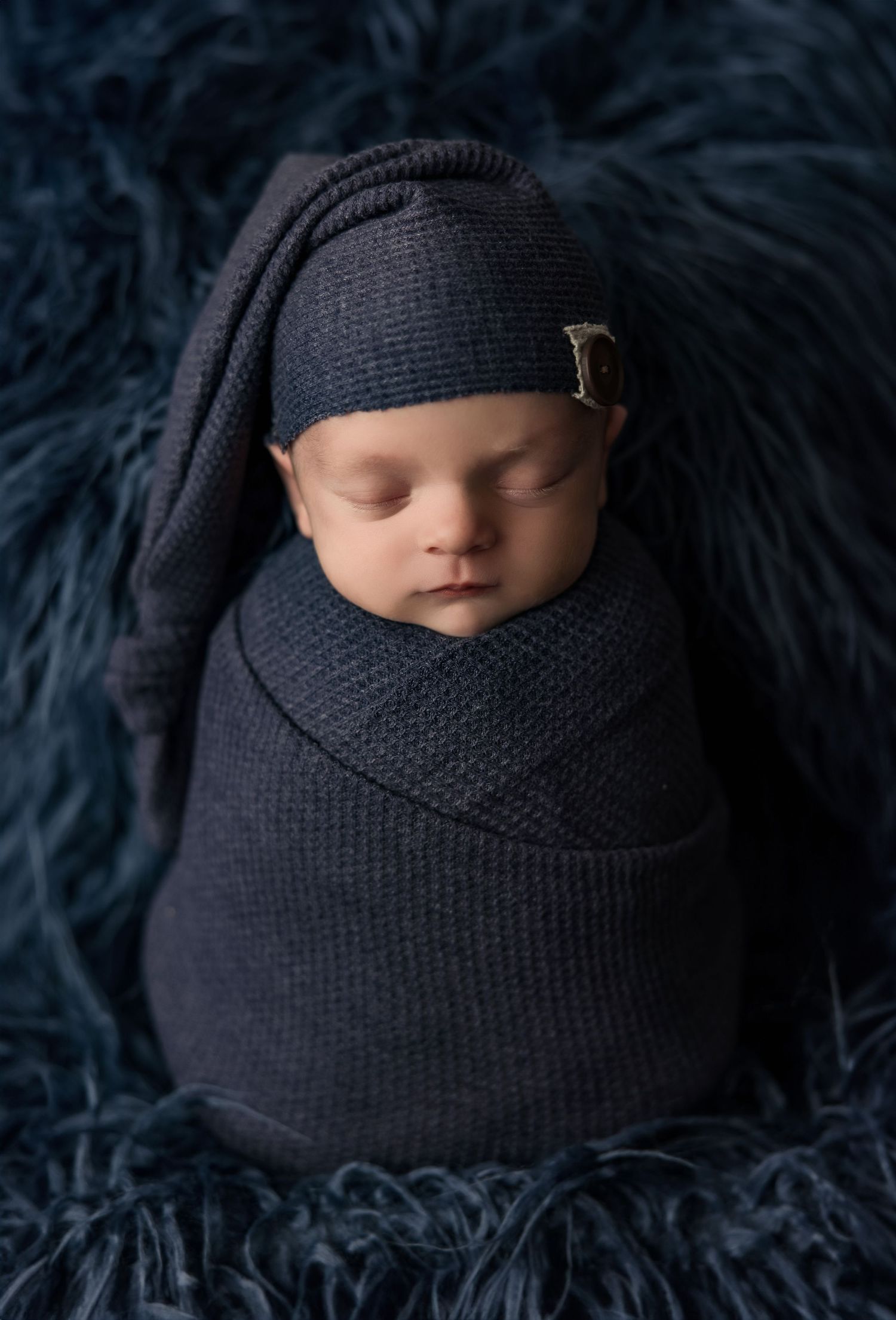 A sleeping newborn baby wrapped in a dark knit blanket against a navy blue furry backdrop.
