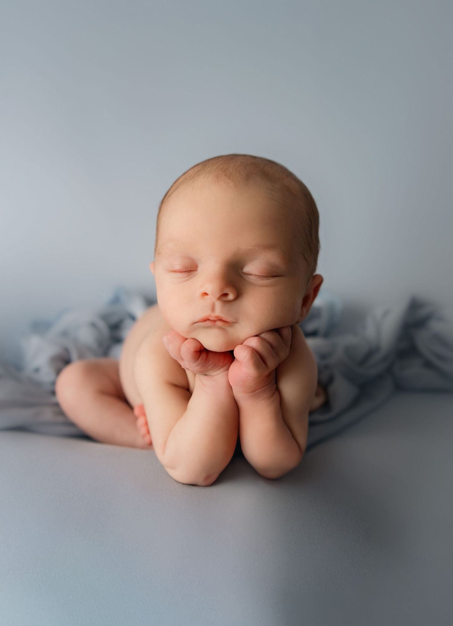 A sleeping newborn baby posed on a gray blanket in froggy position with hands under chin in soft lighting.
