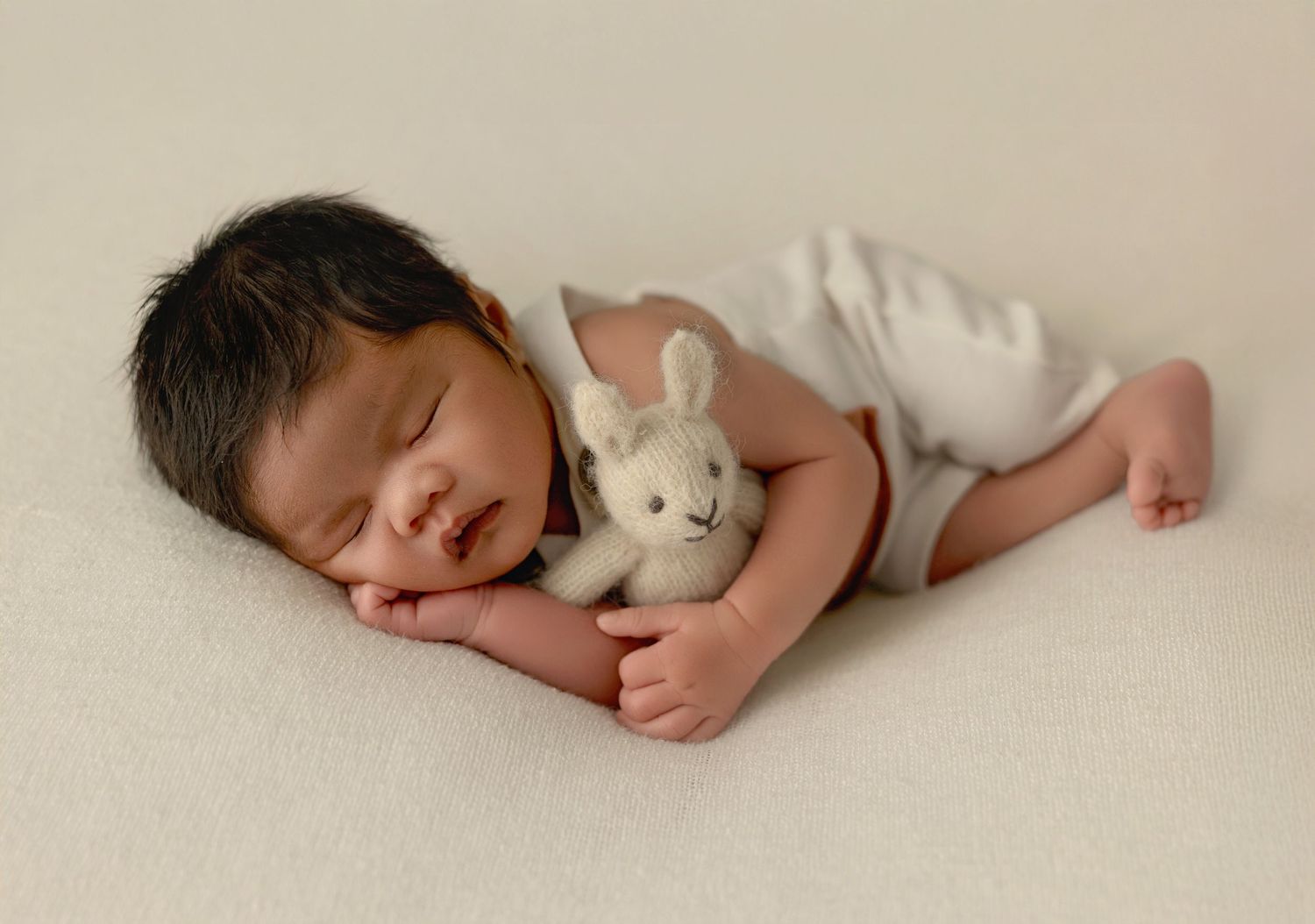 A sleeping baby in white swaddle blanket lies peacefully on a cream colored background.