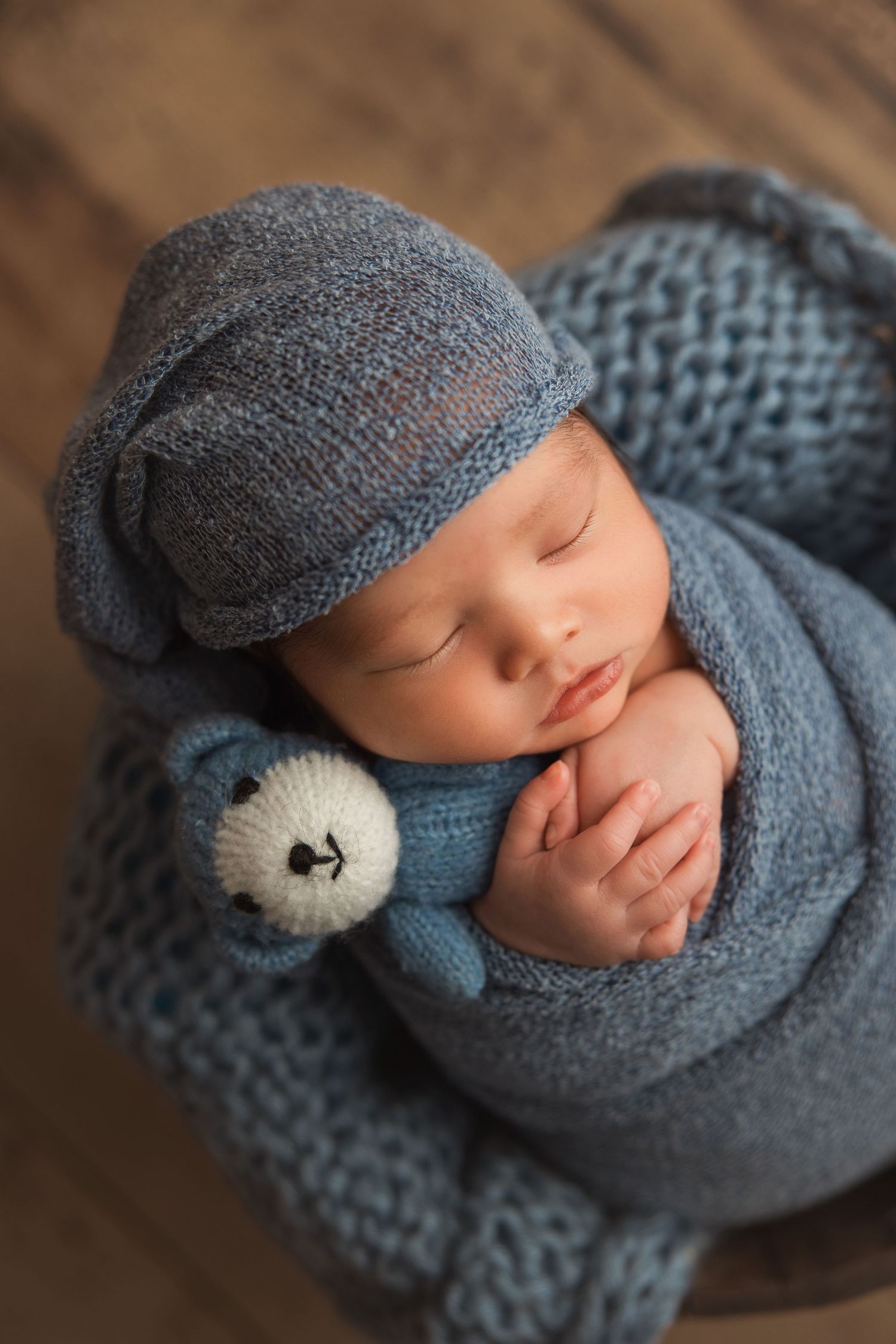 Sleeping baby wrapped in soft gray knit blanket and hat with small teddy bear wearing blue sweater.