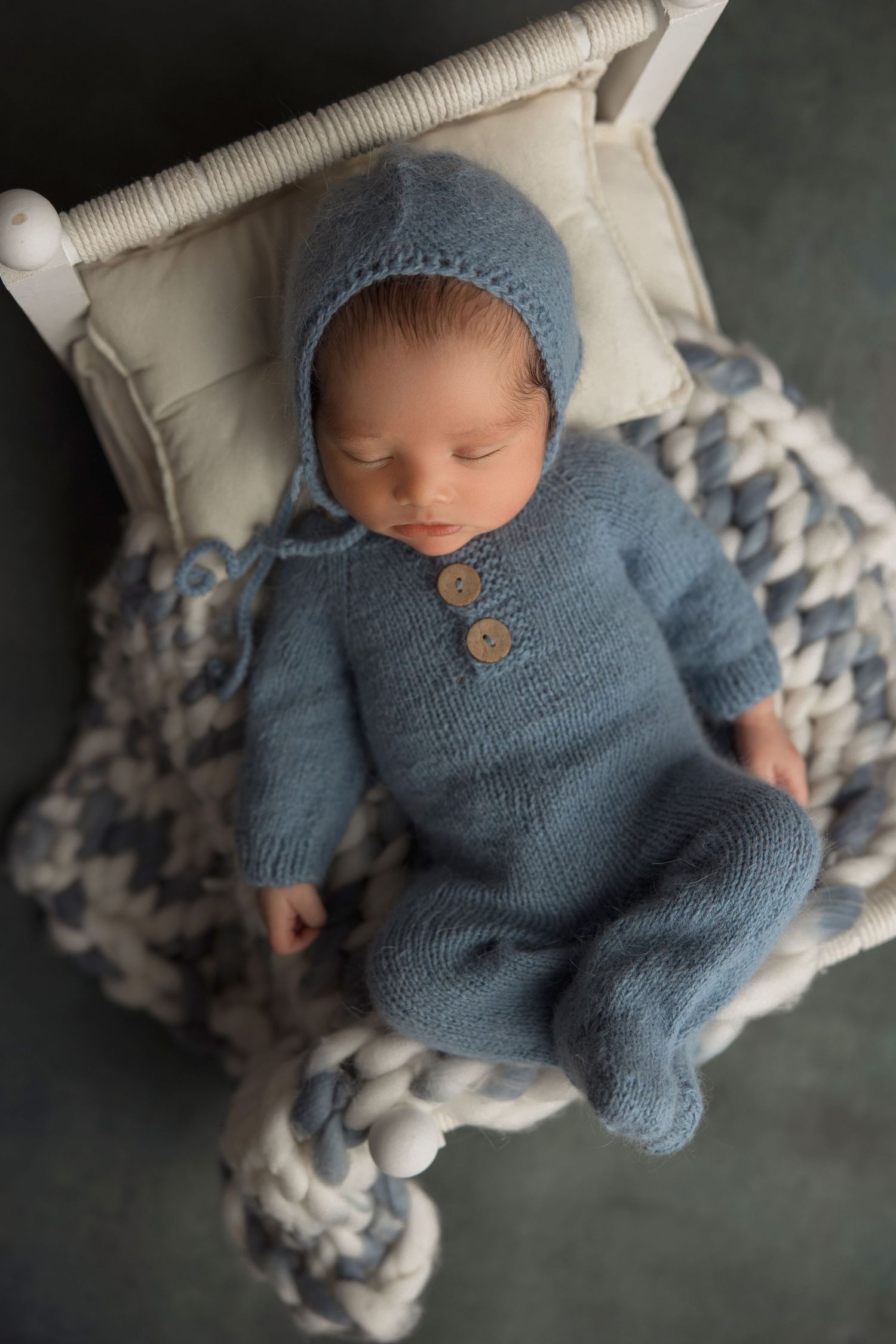 Sleeping baby wearing cozy blue knit sweater and hat rests peacefully on patterned pillow.