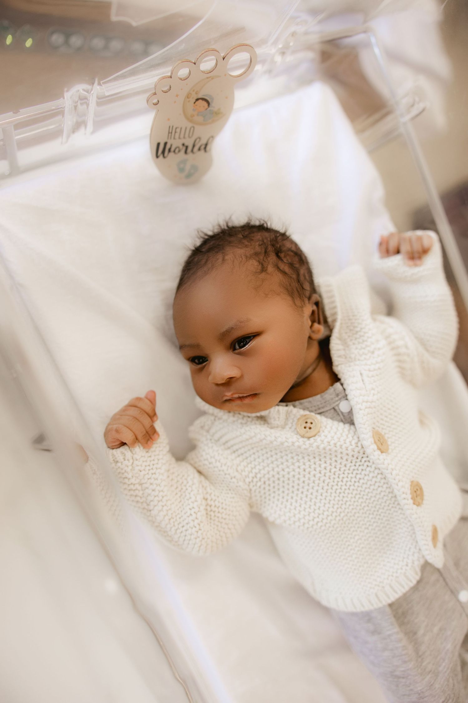 A newborn baby wearing a white knit sweater rests peacefully on white bedding.