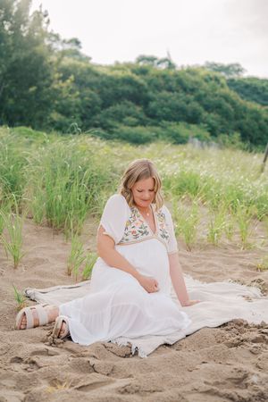 Woman in white dress sitting gracefully on sandy beach surrounded by tall grass and natural scenery.