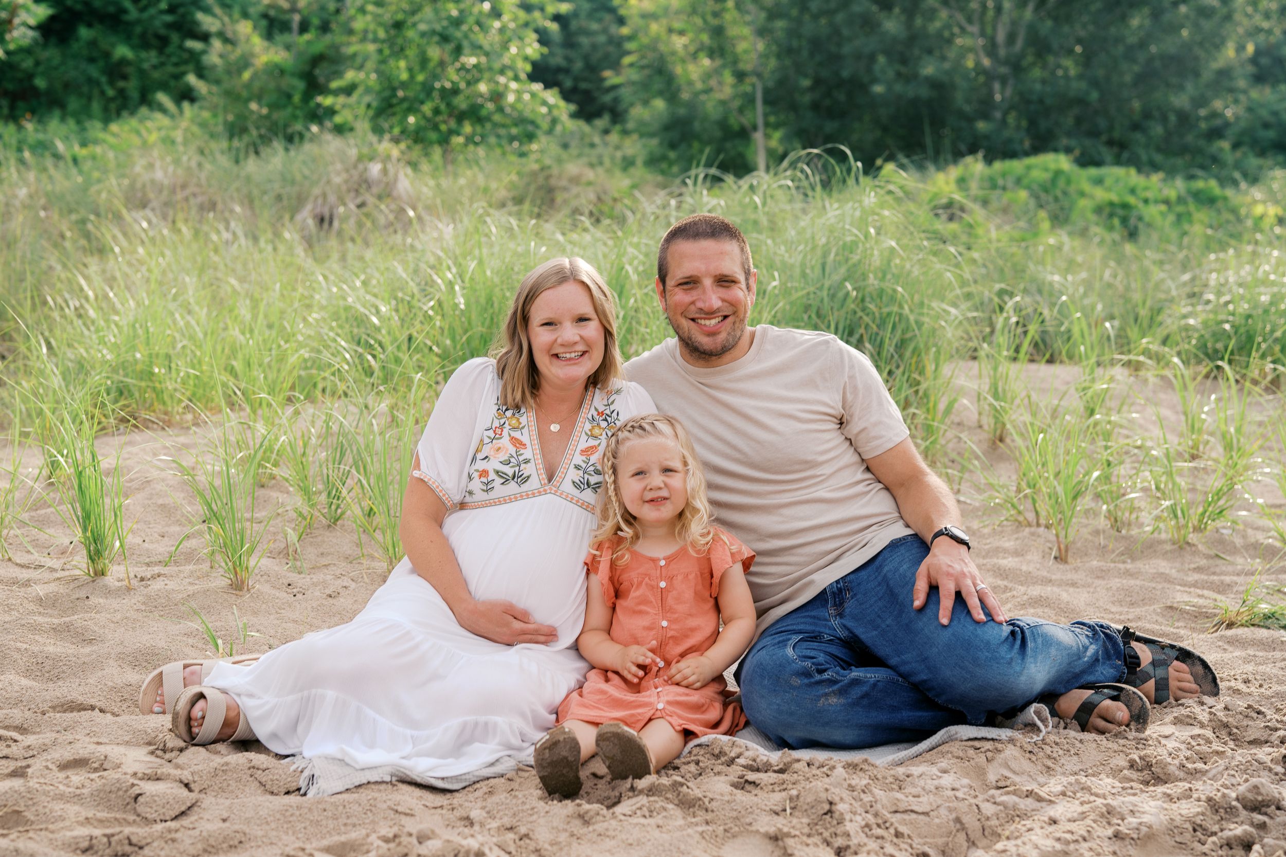 A family sits together on a sandy beach during a maternity photo session on a bright summer day.