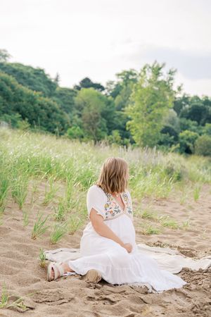 A white dress-clad figure sits peacefully in tall grass on a sandy beach with green hills in background.