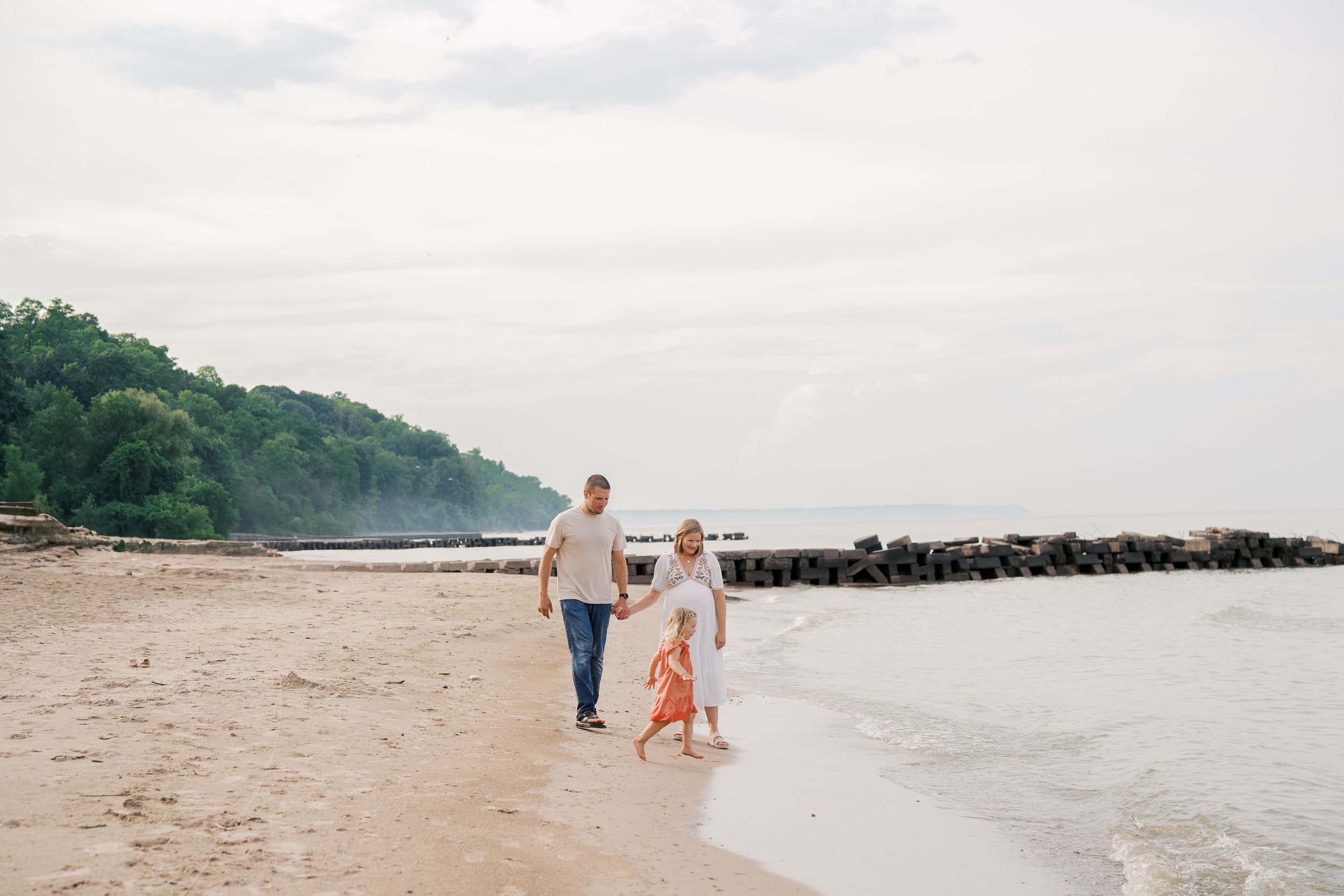 A family walks hand in hand along a beach shoreline with a rocky jetty in the background.