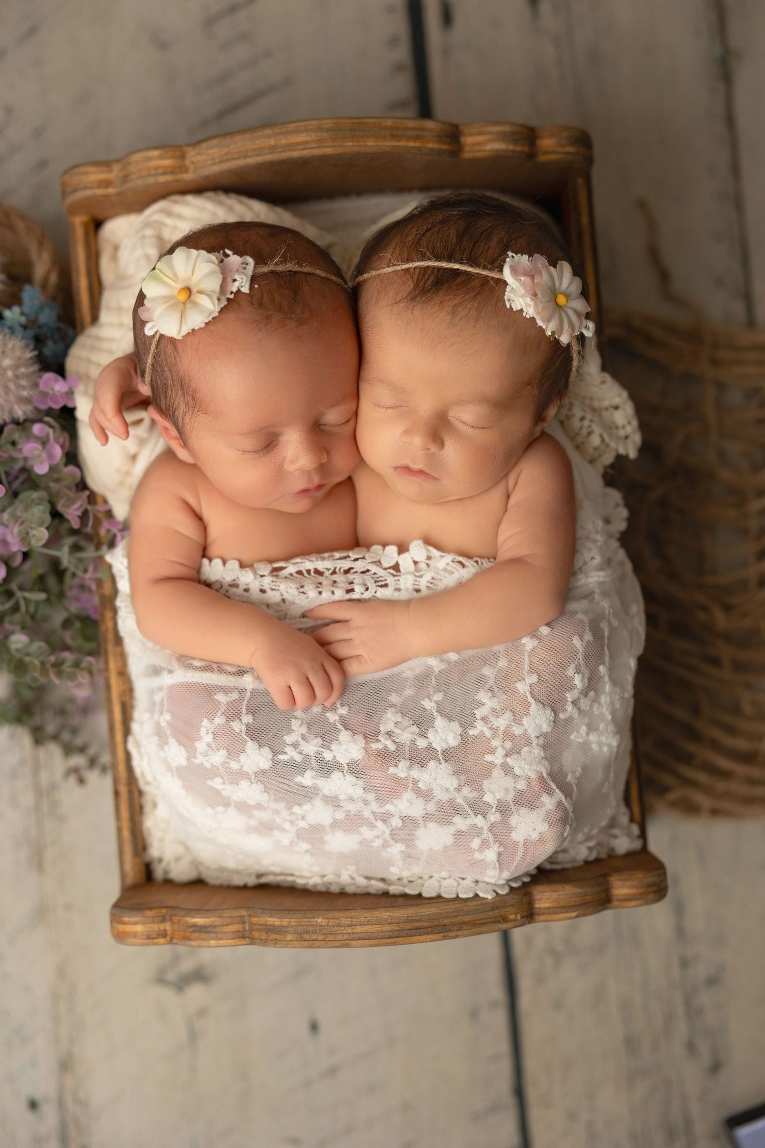 Two sleeping newborns nestled together in a rustic wooden basket with lace bedding and delicate floral decorations.