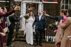 Wedding guests throw pink and white confetti at a joyful celebration outside a brick building.