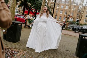 A bride in a flowing white wedding dress twirls on a brick sidewalk while confetti falls around her in an urban setting.