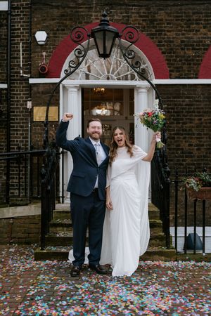 Joyful couple celebrates outside historic brick building with colorful confetti scattered on ground.
