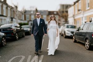A couple walks down a residential street lined with parked cars on their wedding day.