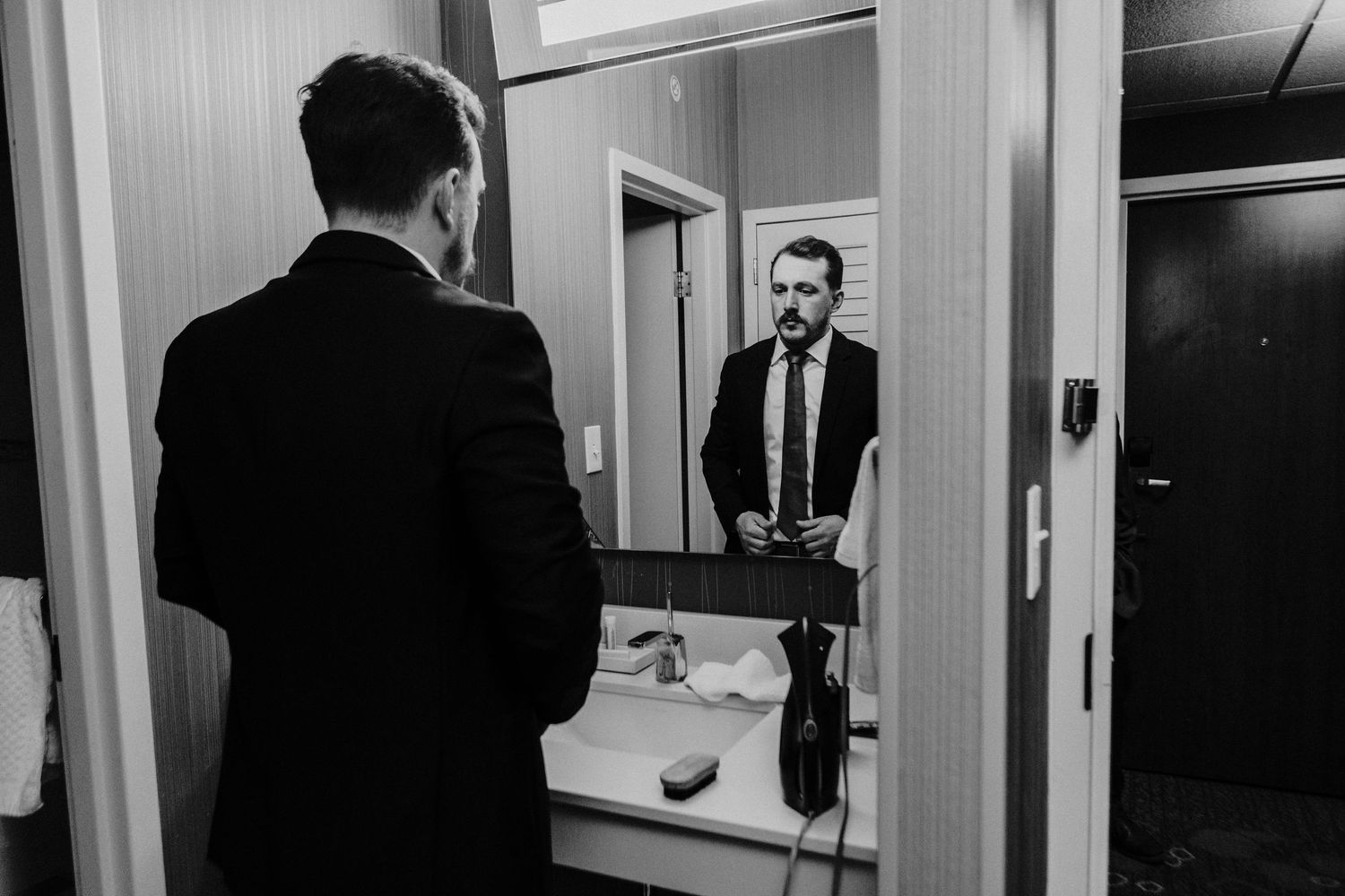 A groom in a black suit adjusts his tie while looking in a hotel bathroom mirror before his wedding.