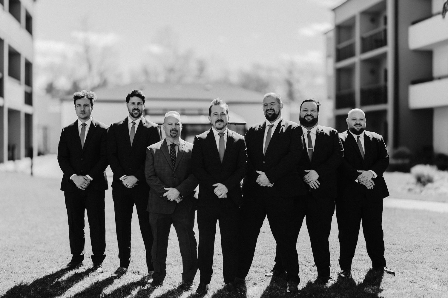 Black and white photo of seven groomsmen in dark suits posing together outdoors at a wedding venue.