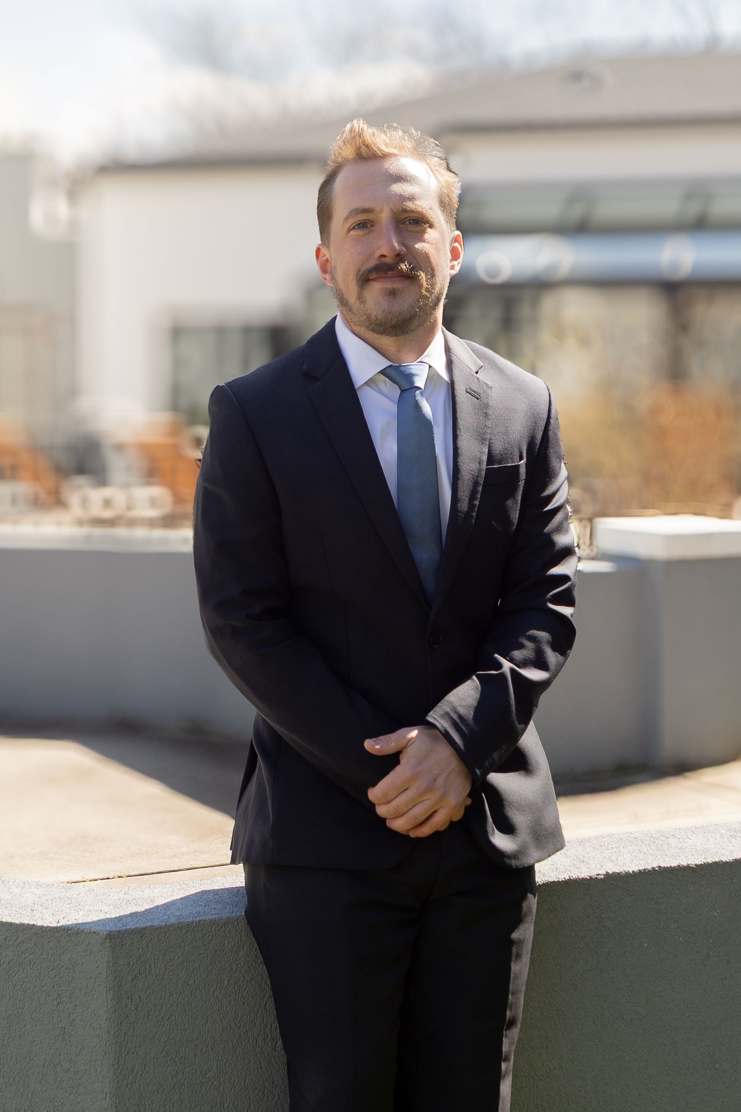 Professional man in dark suit and blue tie posing outdoors on a rooftop terrace on a sunny day.