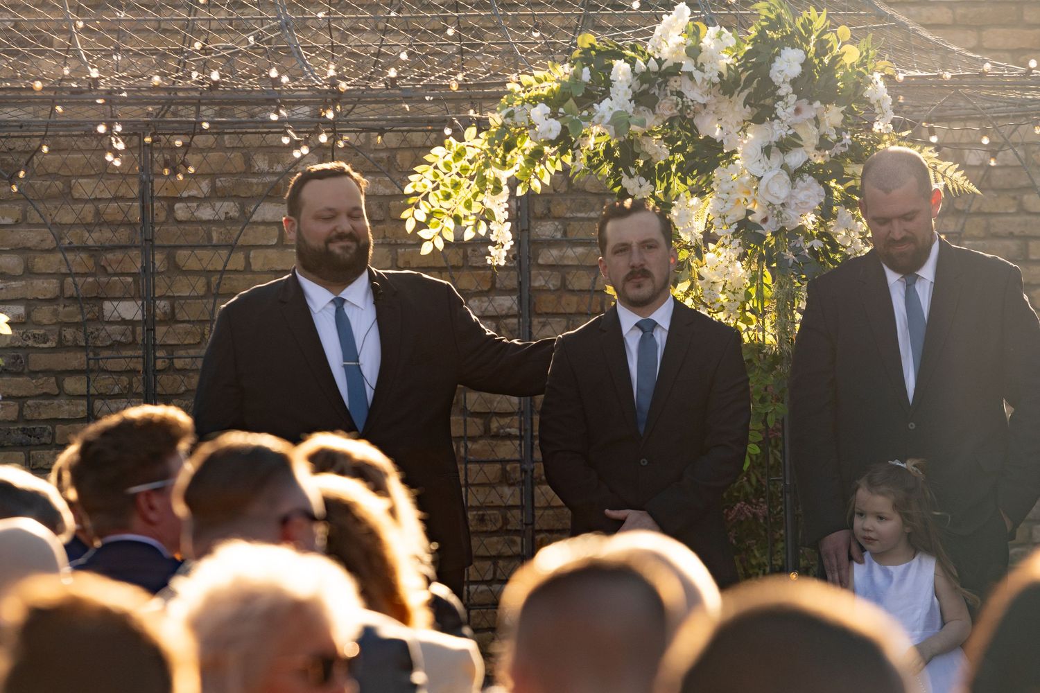 Three groomsmen in dark suits stand at a floral wedding arch outdoors during golden hour ceremony.