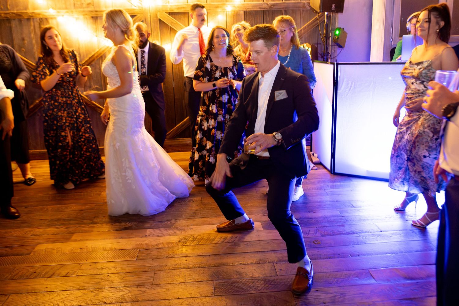 Guests dancing energetically at a wedding reception on a wooden dance floor, with the bride visible in the background.