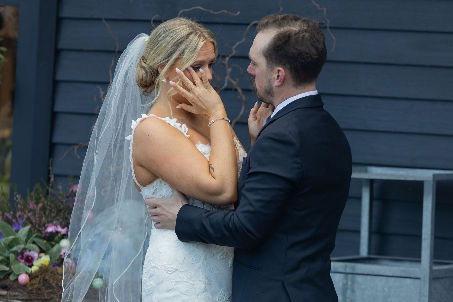 Emotional bride in white lace gown and veil being comforted by groom during outdoor wedding ceremony.