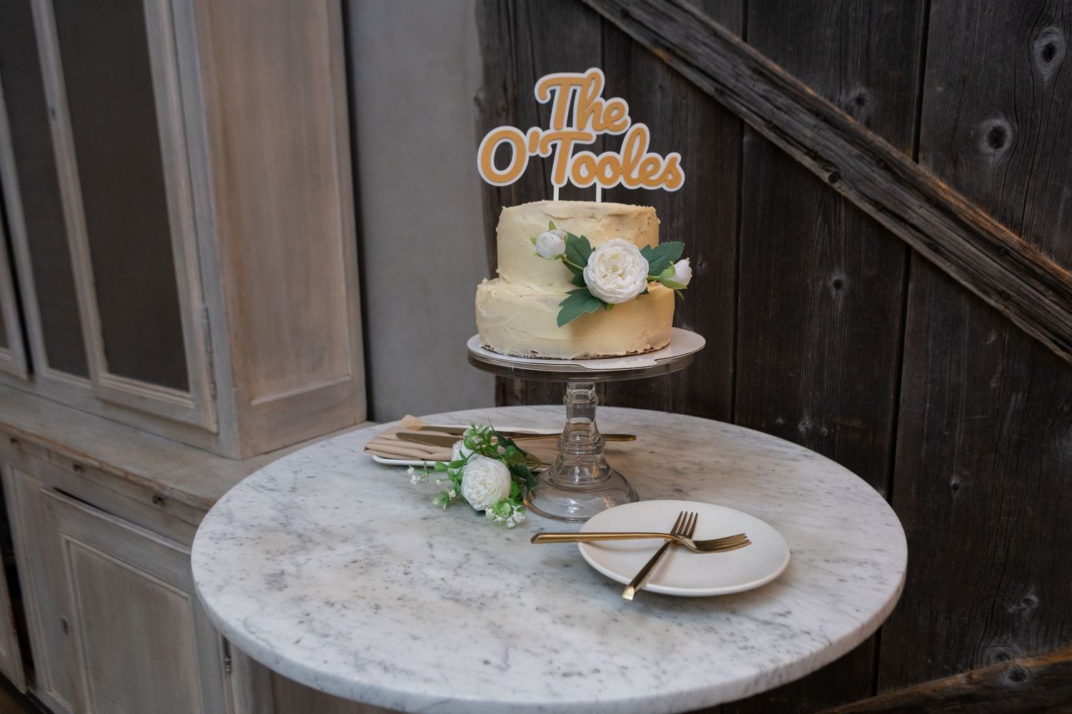 Small two-tier wedding cake with gold The O'Tooles topper on marble table with white roses and wooden utensils.