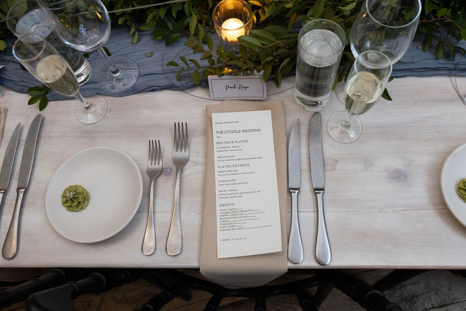 Elegant wedding table setting with white plates, silverware, menu cards, and glowing candles on a rustic wooden table.