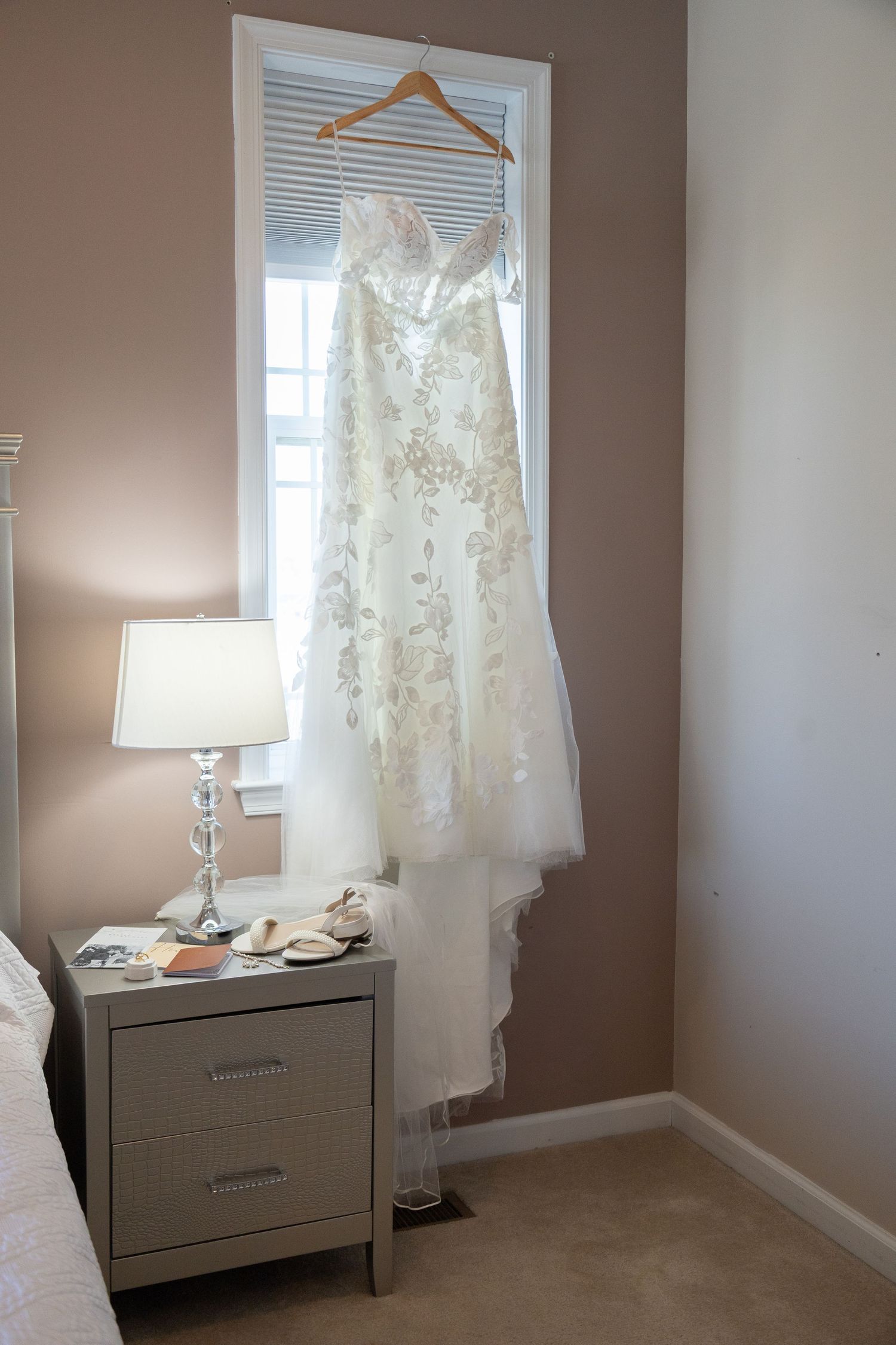 Elegant lace wedding dress hanging on a wooden hanger in a sunlit bedroom window above a white nightstand.