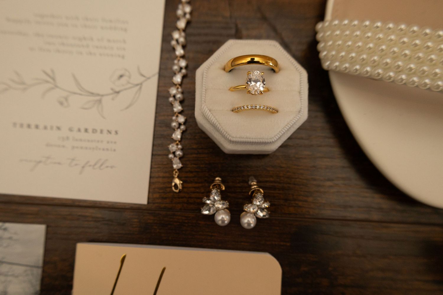 Gold engagement ring with oval diamond in velvet box beside pearl earrings and wedding invitation on wood table.