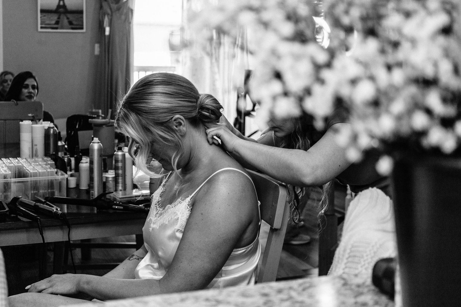 Bride getting hair styled in salon mirror, black and white wedding preparation photo with flowers in foreground.