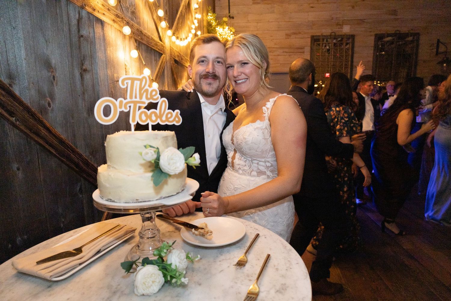 Newlyweds cutting wedding cake at rustic venue, cake topper reads The O'Tooles, bride in white gown.