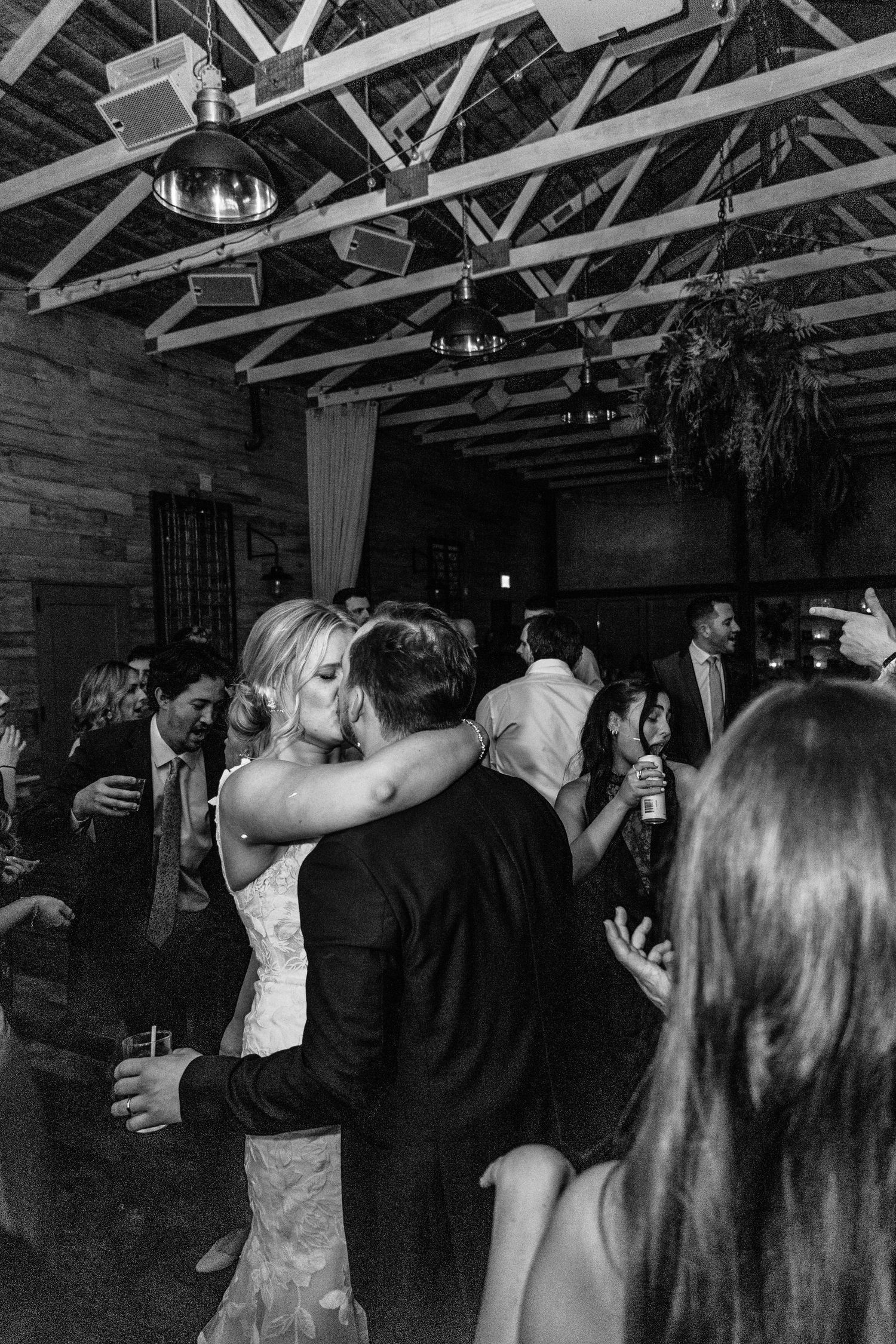 Black and white photo of a couple sharing a kiss on the dance floor surrounded by guests at a wedding reception.
