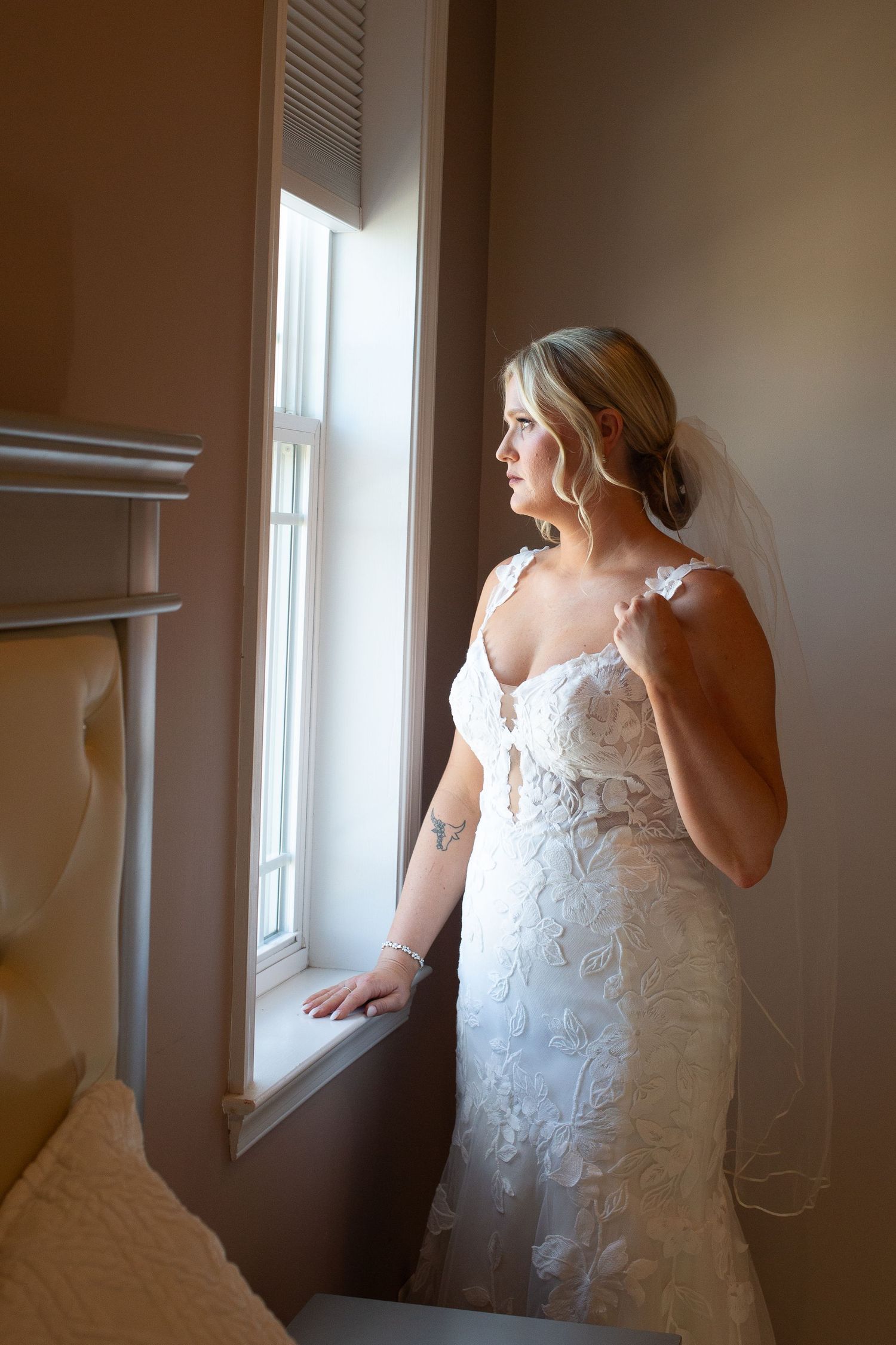 Bride in lace wedding gown and veil gazes out sunlit window, elegant updo, tattoo visible on arm.