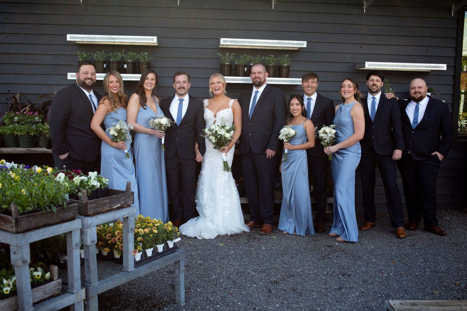 Wedding party posing outdoors in dusty blue bridesmaid dresses and dark suits beside a rustic barn with flower displays.