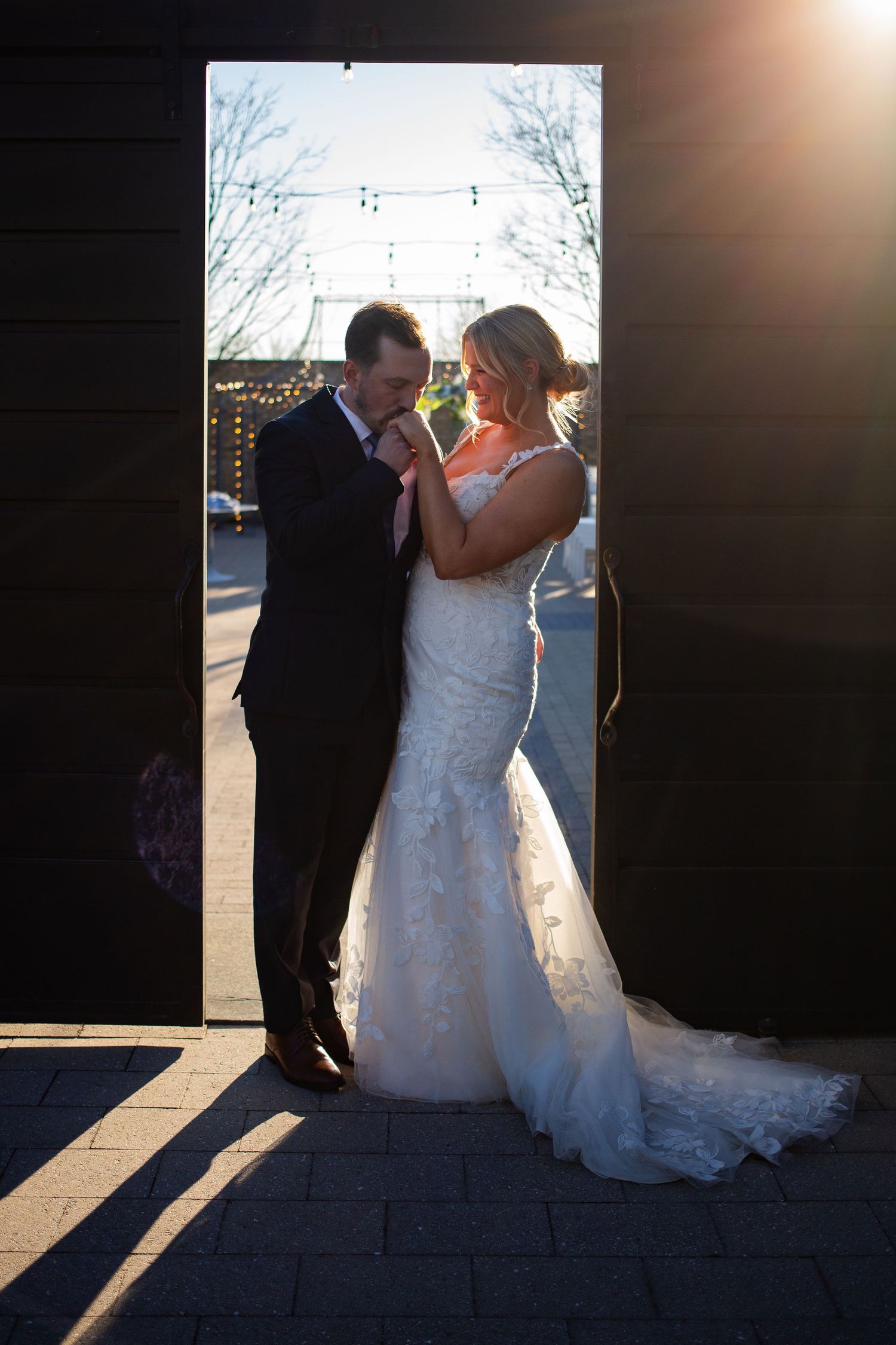 Bride and groom share a romantic moment in golden sunlight framed by dark barn doors in a wedding portrait.