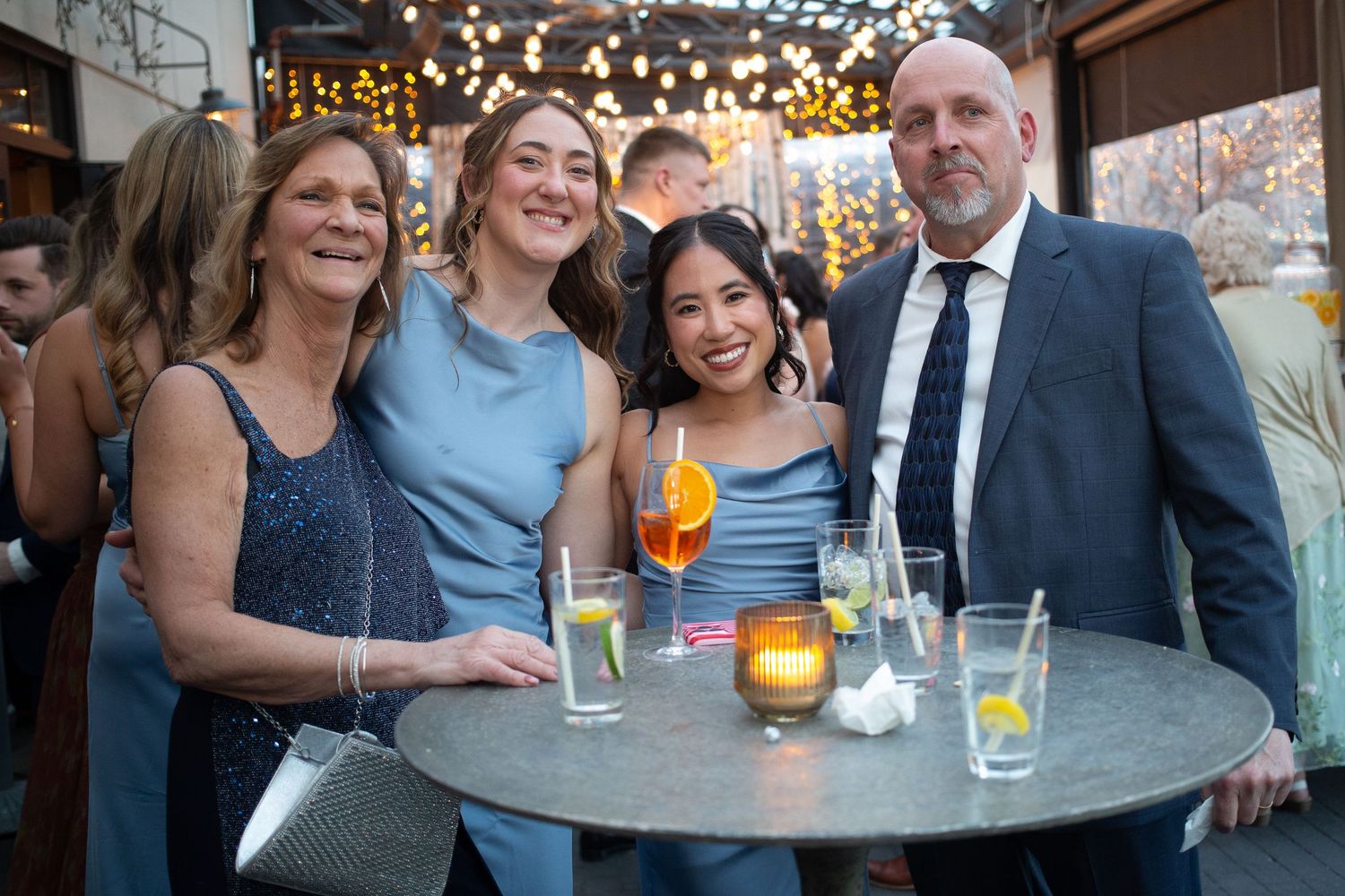Four people smiling together at an outdoor bar event, with cocktails on a round table and string lights in the background.