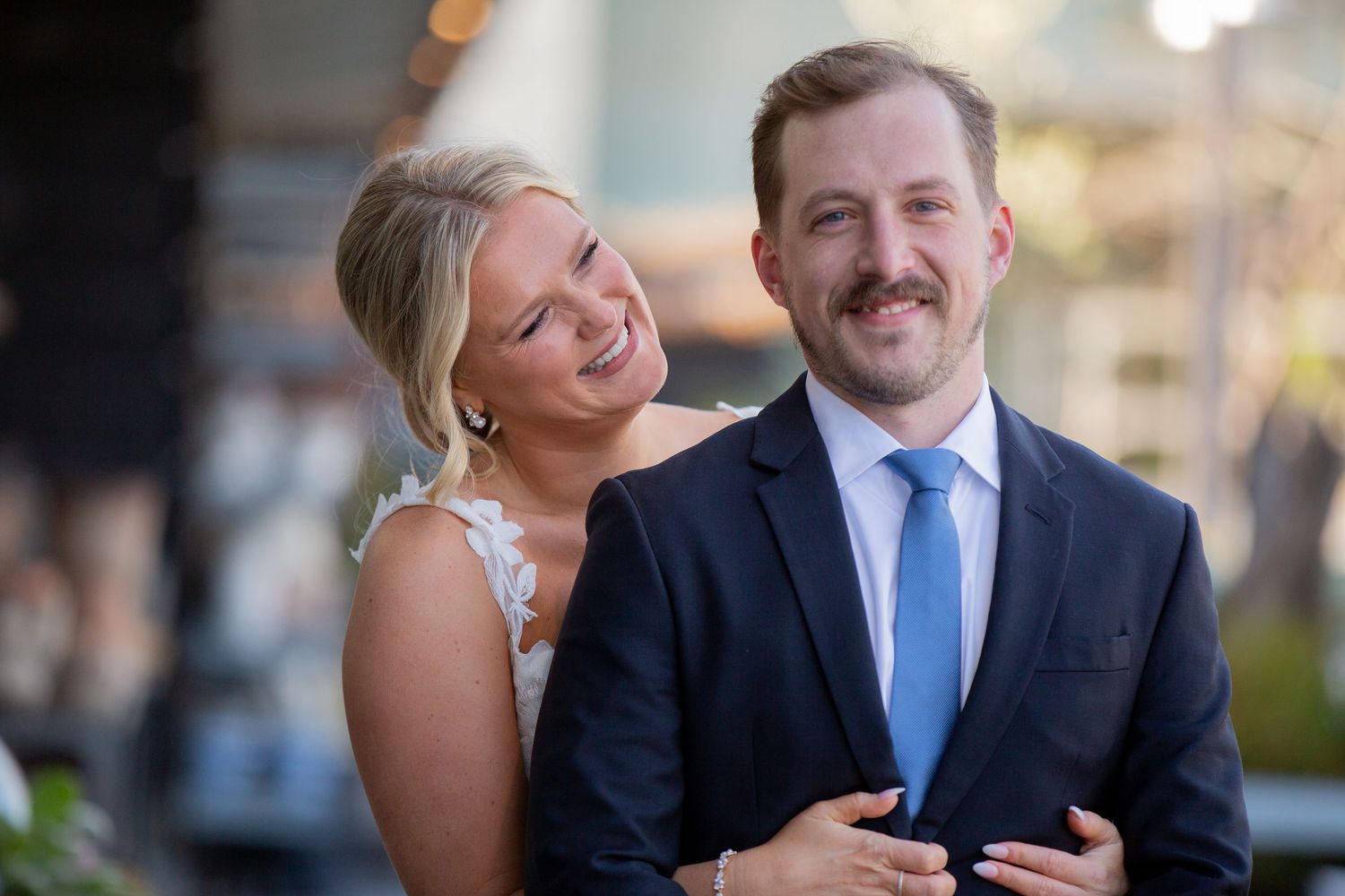 Happy couple posing outdoors, woman in floral dress leaning on man in navy suit with blue tie.