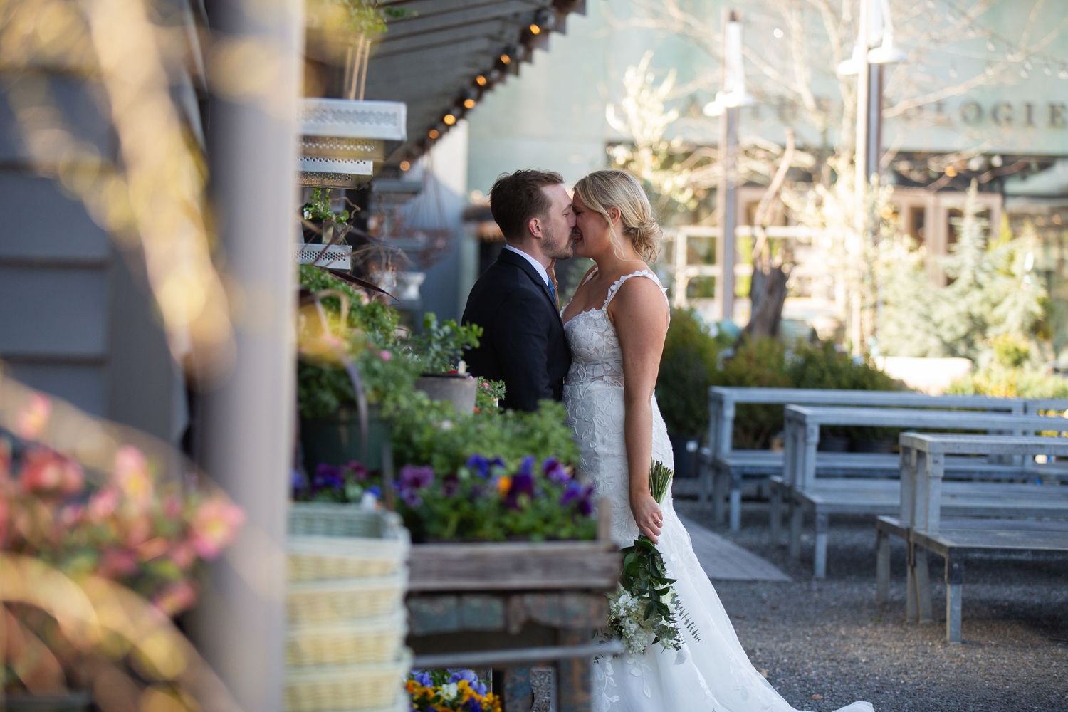 Bride and groom sharing a kiss outdoors at a charming venue surrounded by lush greenery and colorful flowers.